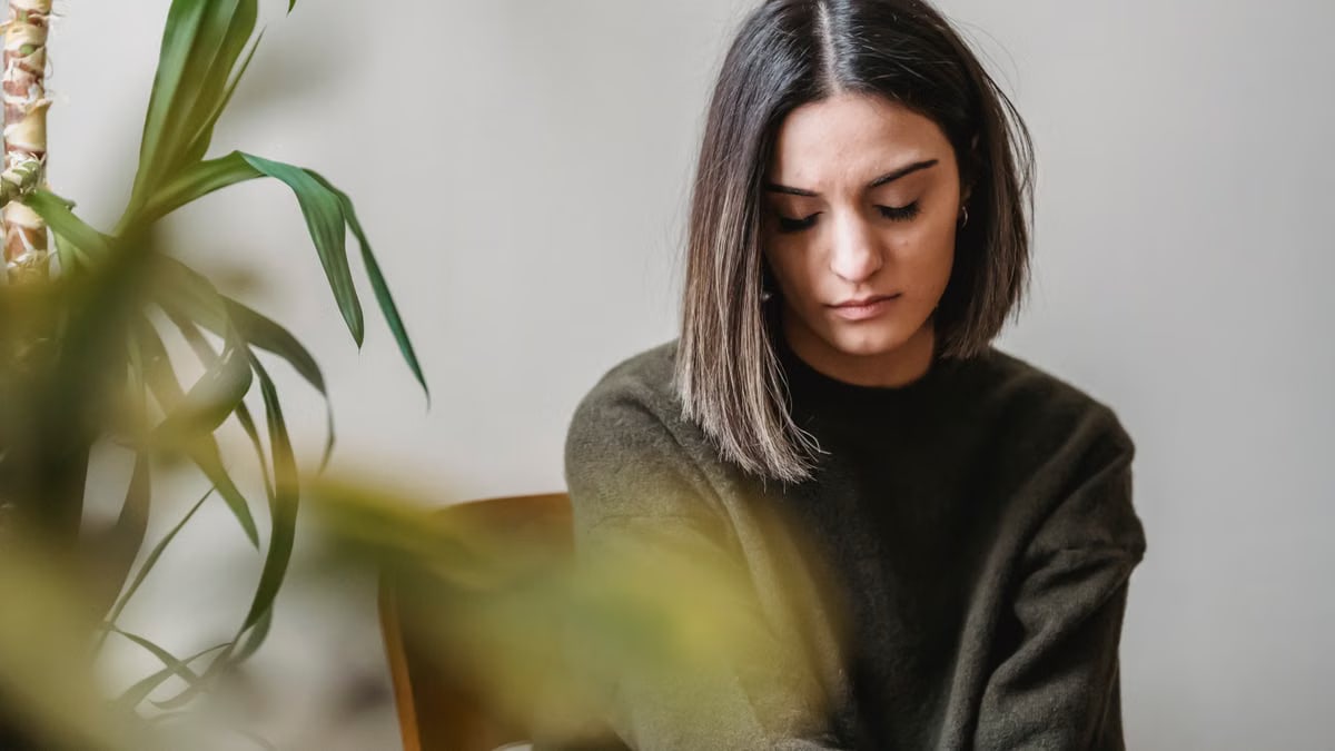 Calm young ethnic lady with dark hair in casual outfit looking down pensively while sitting on chair near potted plant